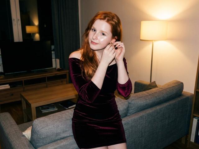 CorrinaGoodland with red hair, wearing a dark purple off-shoulder velvet dress, standing in a room, hands near face, soft smile, glowing lamp in background.