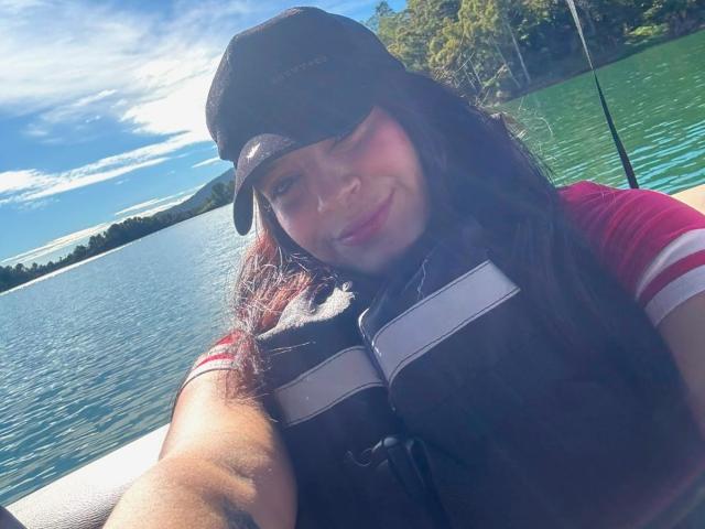 VictoriaFiore with long brown hair, wearing a black cap and red-striped shirt, smiling at the camera, showing her chest in a brown life jacket, on a boat.