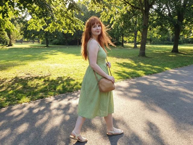 CorrinaGoodland, redheaded, pregnant, green dress, tan sandals, brown crossbody bag, standing on park path, sunlight filtering through trees, looking back, wavy hair, fair skin, soft smile.