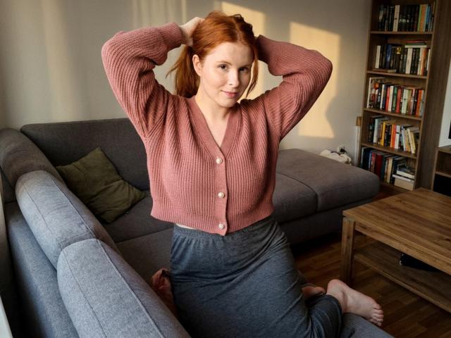 CorrinaGoodland with red hair, pink ribbed cardigan, and grey skirt, hands in hair, sitting on grey couch, wooden coffee table, bookshelf, and sunlight in background.