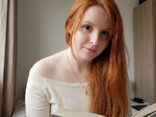 CorrinaGoodland with long red hair, freckles, and pale skin, wearing a white off-shoulder sweater, holding a book, looking at the camera with a slight smile.