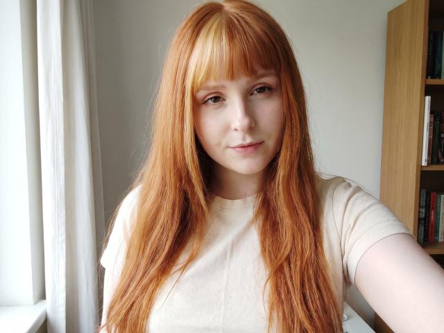 Corrina Goodland, com cabelos ruivos longos, pele clara e seios pequenos, vestindo uma camisa bege, sorrindo levemente, tirando uma selfie perto de uma janela, com uma estante de livros de madeira ao fundo..