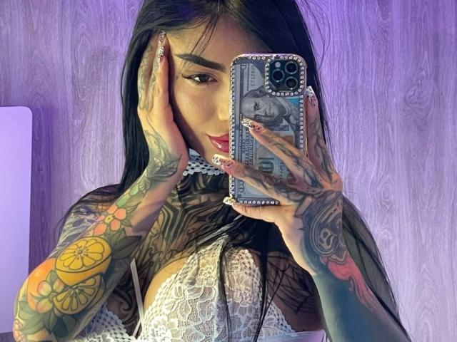 DaniBloome with tattooed arms, holding phone in front of mirror, showing large tits in white lace top, purple background, long black hair, red lipstick, checking reflection.