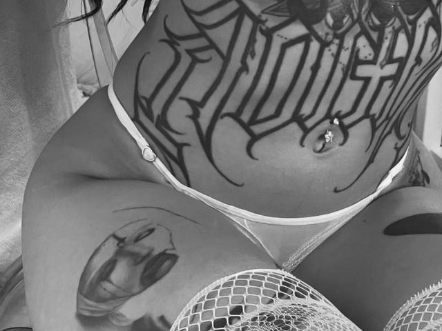 DaniBloome's tattooed midsection with intricate black ink, wearing white fishnet stockings and sheer panties, showing a belly button piercing and tattoos on her thighs. Black and white photo.