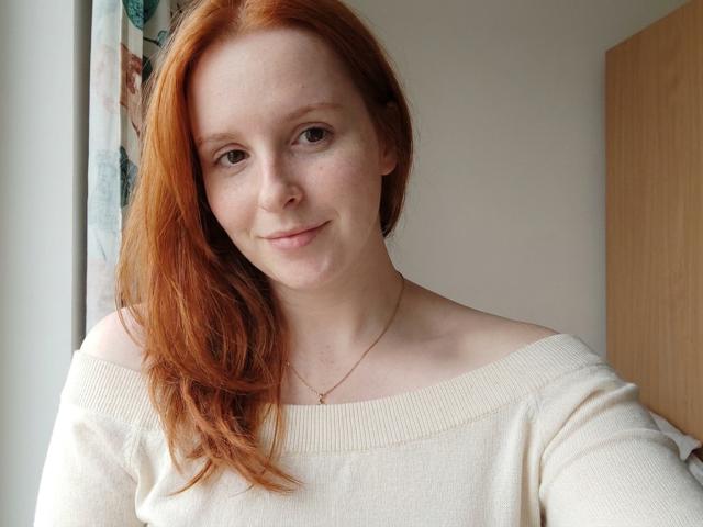 Corrina Goodland ha i capelli rossi, la pelle chiara e delle lentiggini, indossa un maglione bianco con una spalla scoperta, ha un sorriso appena accennato e una collana d'oro, ed è in un ambiente con luce naturale..