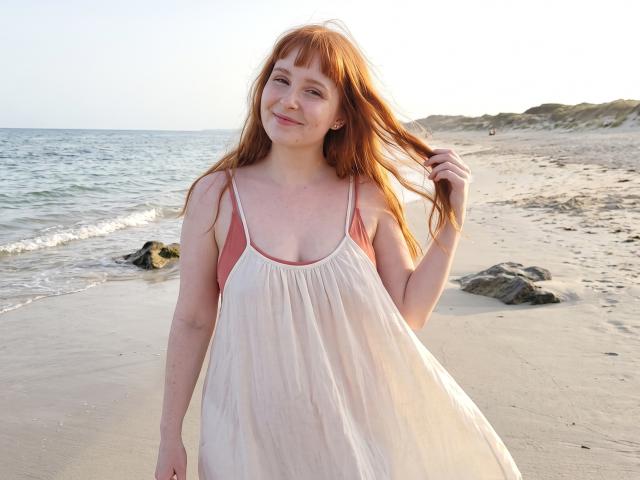 Corrina Goodland, avec des cheveux roux, souriante, portant une robe blanche avec des bretelles roses, se tenant les cheveux, debout sur une plage de sable avec des rochers et l'océan en arrière-plan..