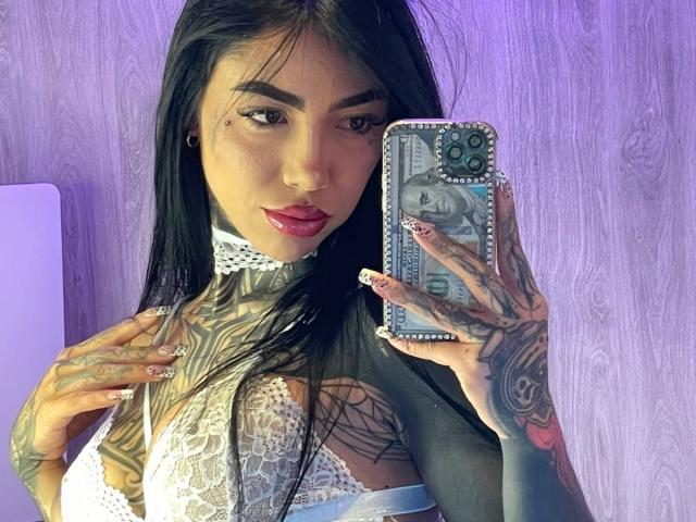 DaniBloome with tattoos, holding phone, wearing lace bra, purple background, long black hair, red lipstick, tattoos on neck, chest, and hands, taking selfie, purple wall behind.