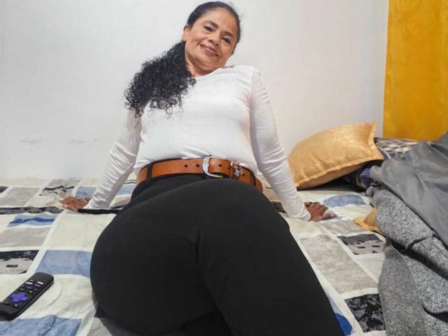 AuroraCandelax, with curly black hair, white long-sleeve shirt, and black pants, sits on a bed, smiling, showing her big ass, with a brown belt and gold pillow.