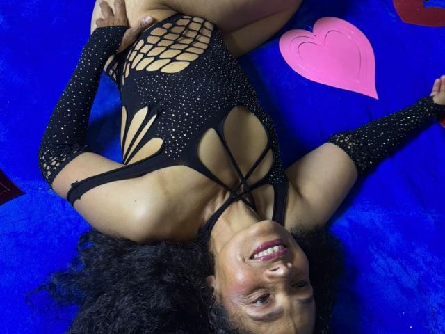 AuroraCandelax with curly black hair, smiling, lying on blue carpet, wearing a black, fishnet-style bodysuit, exposing her tits and showing a big ass. Pink heart sticker nearby.