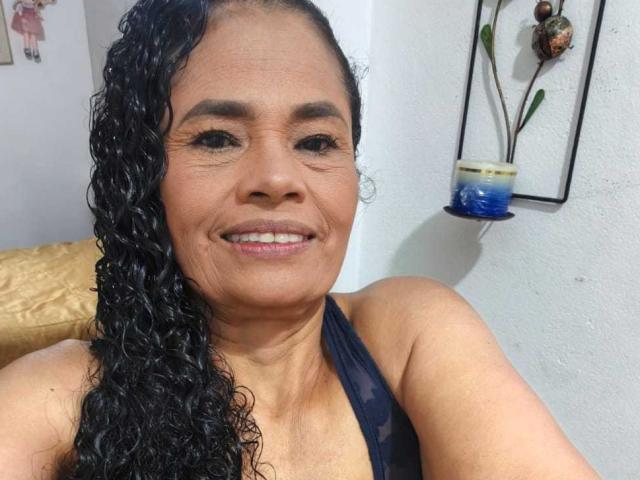 AuroraCandelax has curly black hair, tan skin, and is smiling with pink lipstick, wearing a dark blue halter top, showing her small tits and ass in a selfie.