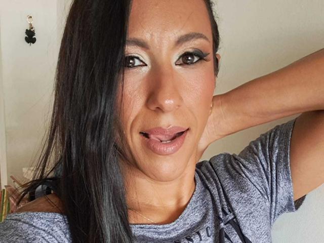 Jade Foster tem a pele morena, cabelos longos e pretos, e está vestindo uma camisa cinza. Seus lábios estão ligeiramente entreabertos, mostrando um pequeno pedaço de sua língua. Ela tem um delineador marcante..