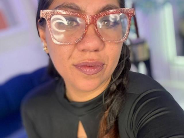 SilvanaLane with brown skin and long braided hair, wearing red glittery glasses with reflective eyes, and a black shirt, showing pouty lips.
