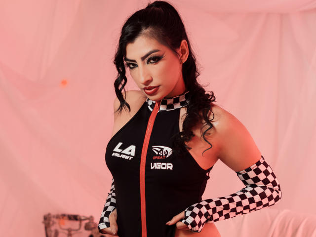 NinnaPiers with black wavy hair, wearing a black and checkered sleeveless top, exposing her tits, with a zipper, and matching gloves, against a pink background.