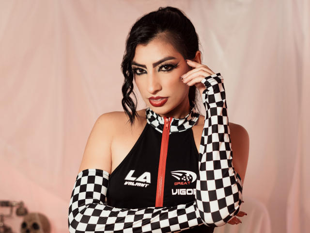 NinnaPiers with dark hair in a ponytail, wearing a black and white checkered top with a red zipper, and matching checkered sleeves, has her hand on her face.