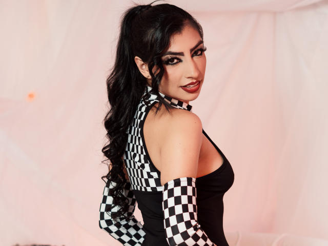 NinnaPiers with long black hair in a ponytail, wearing a black and checkered sleeveless top, showing her bare shoulders and back, smiling at the camera.