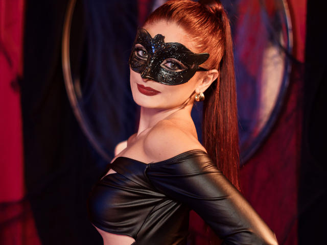 JessieDiaz with red hair in a ponytail, wearing a black sparkly mask, black off-shoulder leather top, and gold earrings, showing her tits and looking back with red lipstick.