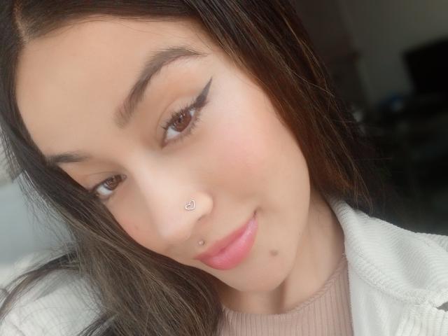 LaurenTyler with brown eyes, black eyebrows, heart-shaped nose stud, nose ring, pink lips, subtle smile, long brown hair, white jacket, and pink ribbed top, looking slightly down.