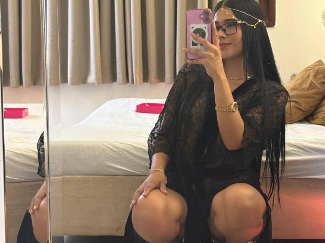 AntonellaMonroe with long black hair, glasses, gold headpiece, black lace robe, squatting, holding pink phone, visible pussy through robe, mirror reflection, gold watch, beige curtains.