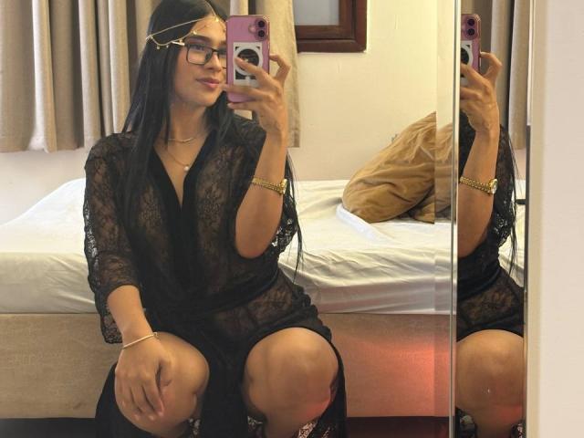 AntonellaMonroe, with long black hair and glasses, wearing a sheer black robe, squatting, holding a phone, showing her ass, in a mirror selfie. Bedroom setting, gold headband.