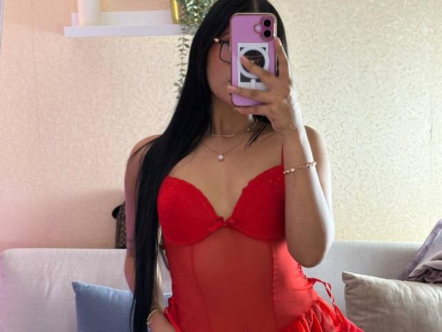 AntonellaMonroe with long black hair, wearing a red lace babydoll, holding a pink phone, covering her face, showing her tits, in a cozy room with a white couch.