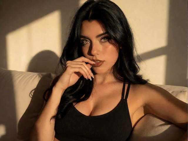 ZoeeMar, con cabello largo y negro, vestida con un top deportivo negro, sentada en un sofá, con la luz del sol proyectando sombras, con la mano cerca de sus labios, mirando seductoramente a la cámara..