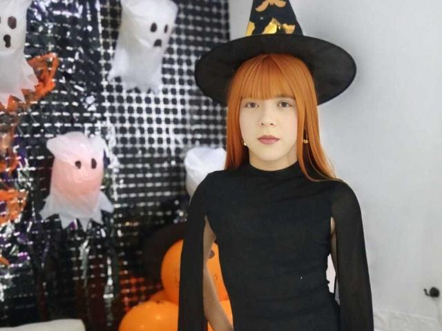 AndrewTaylor69, una chica pelirroja atractiva vestida con un disfraz de bruja negro, con un sombrero puntiagudo, mangas transparentes y una blusa ajustada, está de pie con una expresión neutra, sin mostrar senos ni glúteos visibles..