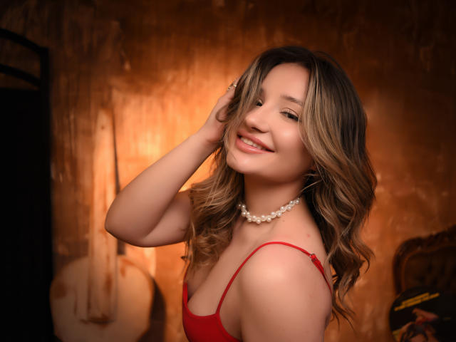 EvaBlazze with wavy blonde hair, red dress, pearl necklace, smiling, touching her hair, showing her tits, soft light, warm brown wall, acoustic guitar in the background.