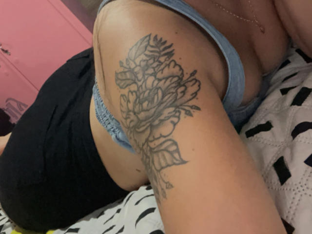SarahGood's tattooed arm with a detailed black rose and leaves on her upper arm, wearing a blue bra and black shorts, lying on a patterned bed.