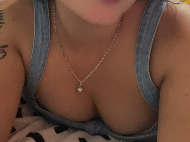 SarahGood's tits in a blue denim top, showing cleavage, with a silver chain necklace and a small heart pendant, her face partially blurred, and a tattoo on her arm.