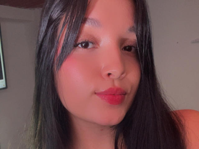 SarahGood has dark straight hair, red lipstick, and a slight smile. Her nose is small and she has a subtle pinkish blush on her cheeks.