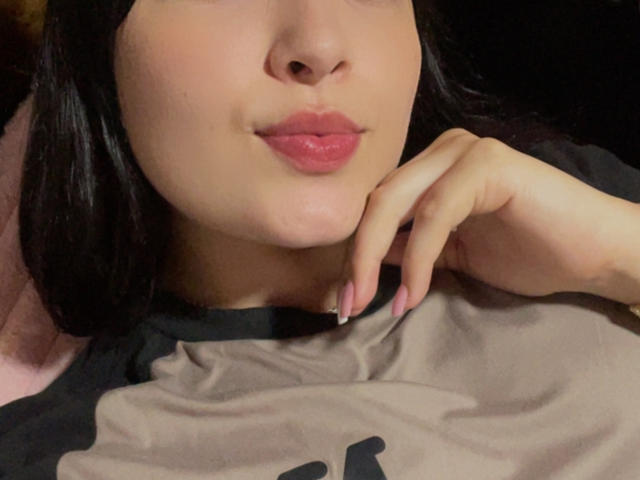 SarahGood's face, with pink glossy lips and dark hair, rests on a black and gray shirt. Her right hand, with manicured nails, touches her chin.