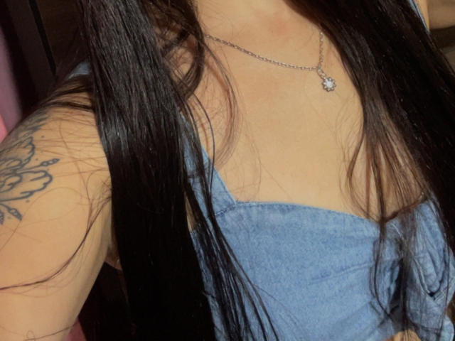SarahGood's chest, showing her tits in a blue top, with long black hair, a tattoo on her right arm, and a silver necklace with a small flower pendant.