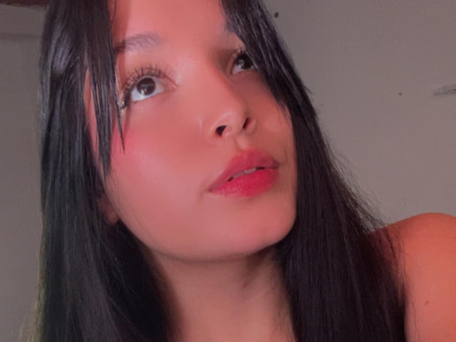 SarahGood with dark straight hair, red lipstick, and light skin, looking to the side with blue eyes and slightly parted pink lips, in a selfie-style image.