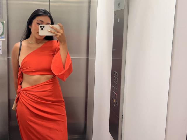 AmeliRoux in an elevator wearing a red one-shoulder dress with a cutout, holding a phone, showing her tits and ass, long black hair, mirror selfie.
