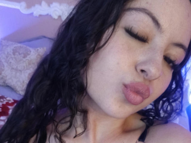 MiiaBennet with curly black hair, closed eyes, and pouty pink lips, wearing a black strap, has a small nose ring, and a light skin tone.