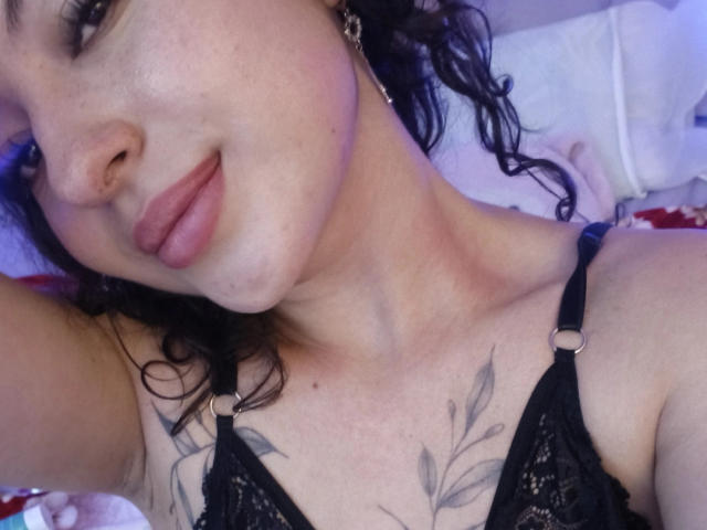 MiiaBennet with curly black hair, wearing black lace bra, showing tattooed chest, pink lips, slight smile, and gold earrings, taking a selfie. Boobs visible, upper body.