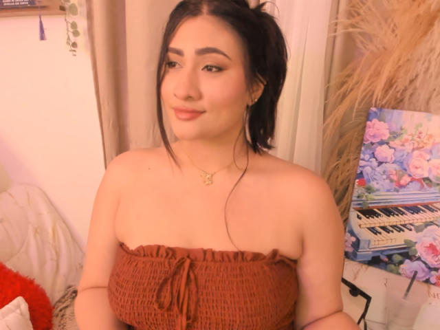 ElyRoss with dark hair in a ponytail, brown strapless top, light skin, subtle makeup, small nose ring, and floral necklace, standing near a piano with flower decals.