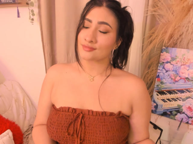 ElyRoss with dark ponytail, closed eyes, and subtle smile, wearing a brown, strapless, textured top, showing her tits, in a cozy, warmly lit room with a keyboard.