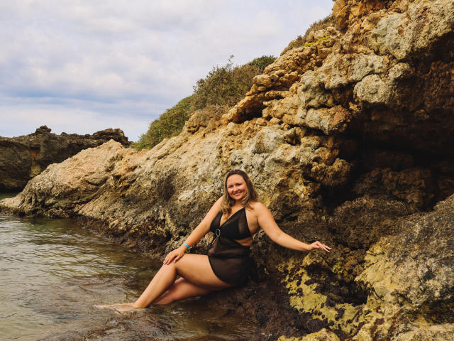 SallyVerda with blonde hair, sitting on rocky shore, wearing black sheer dress, showing her ass, smiling, legs in water, hand on rock, cloudy sky above.
