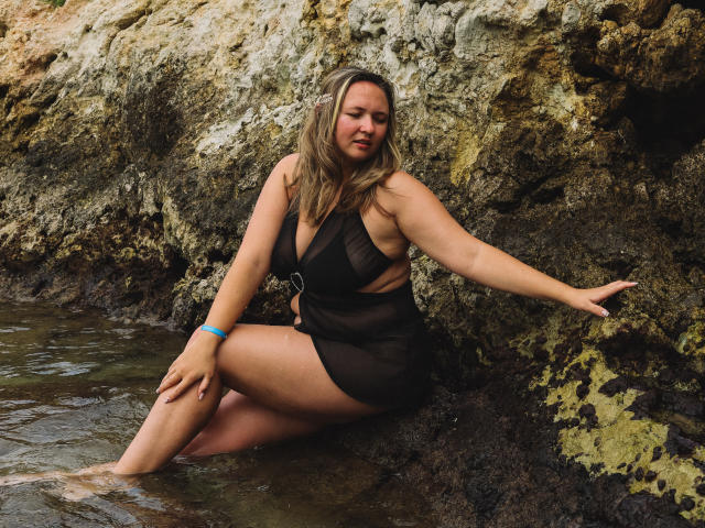 SallyVerda with tanned skin, brown hair, and medium-sized tits, sits in shallow water wearing a sheer black dress, arm on rocky wall, blue bracelet, looking slightly annoyed.