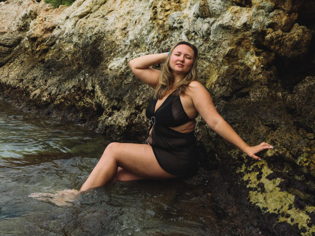 SallyVerda with tits in a black sheer dress, sitting in rocky water, hand on head, eyes closed, legs in water, rocks behind her, blonde hair, slightly wet.