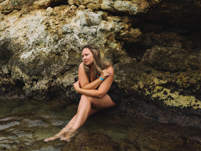 SallyVerda, with blonde hair, sits against rocky shore, legs in clear water, arms crossed over chest, wearing black swimsuit, looking to side, water droplets on skin.