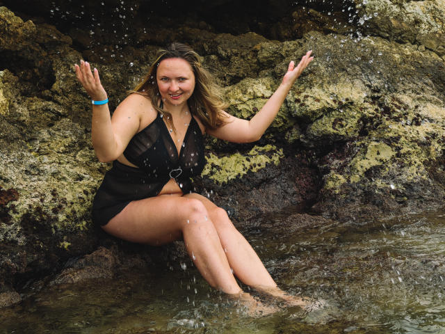 SallyVerda with wet black dress, sitting on rocky shore, legs in water, hands raised, smiling, blonde hair, blue bracelet, water splashing, medium-sized tits, relaxed, natural look.
