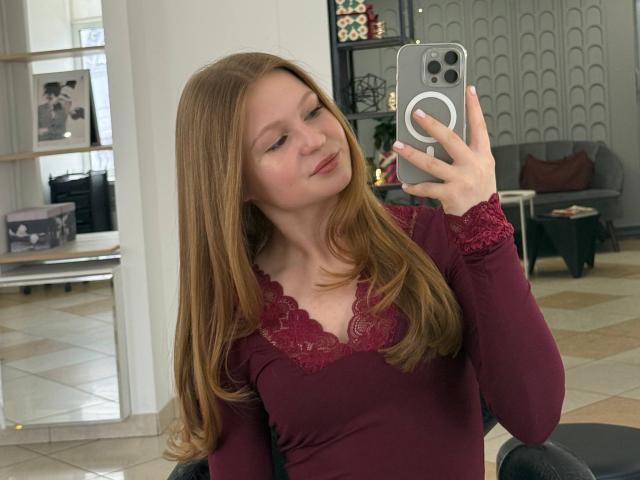 AkilahGhil with long red hair, wearing a maroon lace-top long-sleeve shirt, taking a mirror selfie with a smartphone, showing a slight smile and relaxed expression.