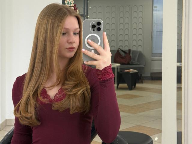AkilahGhil with long, straight, red-brown hair, wearing a maroon lace-trimmed top, holding a silver iPhone, taking a mirror selfie, showing a serious, slightly pouty expression.