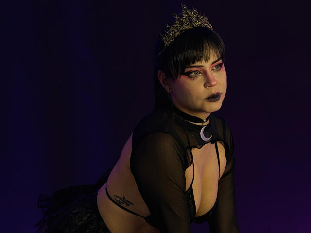 MadameDesadex with black mesh top, crescent moon choker, gold crown, dark lipstick, pink eyeshadow, black bob, tattoo on right side, kneeling, purple-blue lighting, showing her tits.