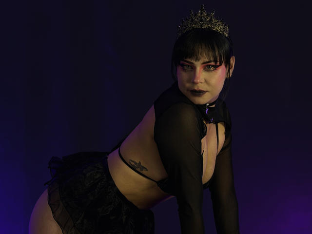 MadameDesadex with black hair, goth makeup, crown, sheer black top, black skirt, and collar, leaning forward, showing her tattooed side and black lipstick. Dark, purple-lit background.