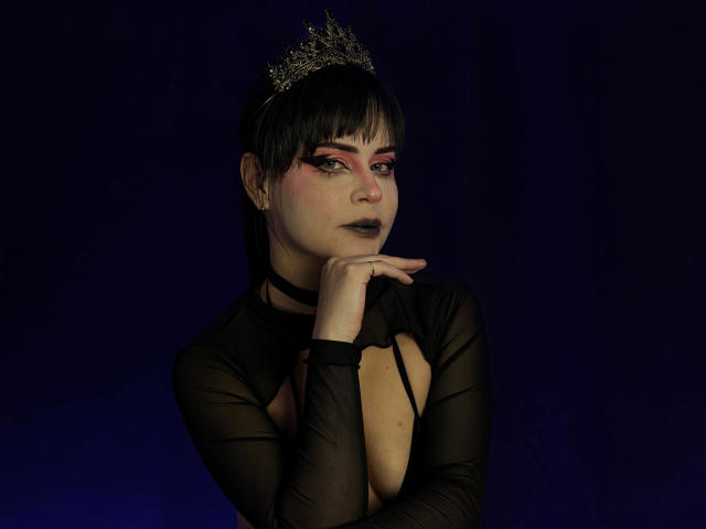 MadameDesadex with dark makeup, black sheer top showing tits, choker, and crown, hand under chin, dark hair, pink and black eye makeup, against dark background.