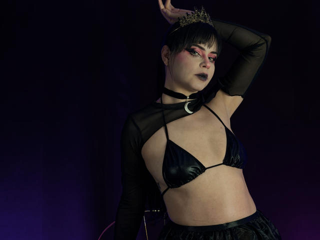 MadameDesadex with black bra, sheer sleeves, crown, dark lipstick, and makeup, arm raised, black choker, purple lighting, black hair, and a sexy, confident, goth-style look.