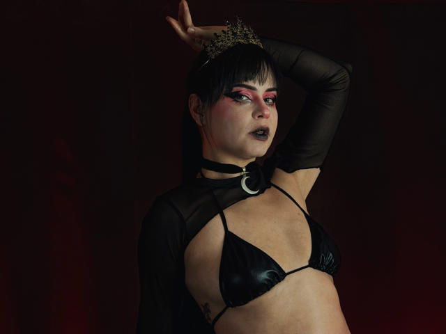 MadameDesadex with black bra, shiny tits, black makeup, pink eyeshadow, choker with crescent moon, black long sleeves, crown of small golden flowers, dark red background.